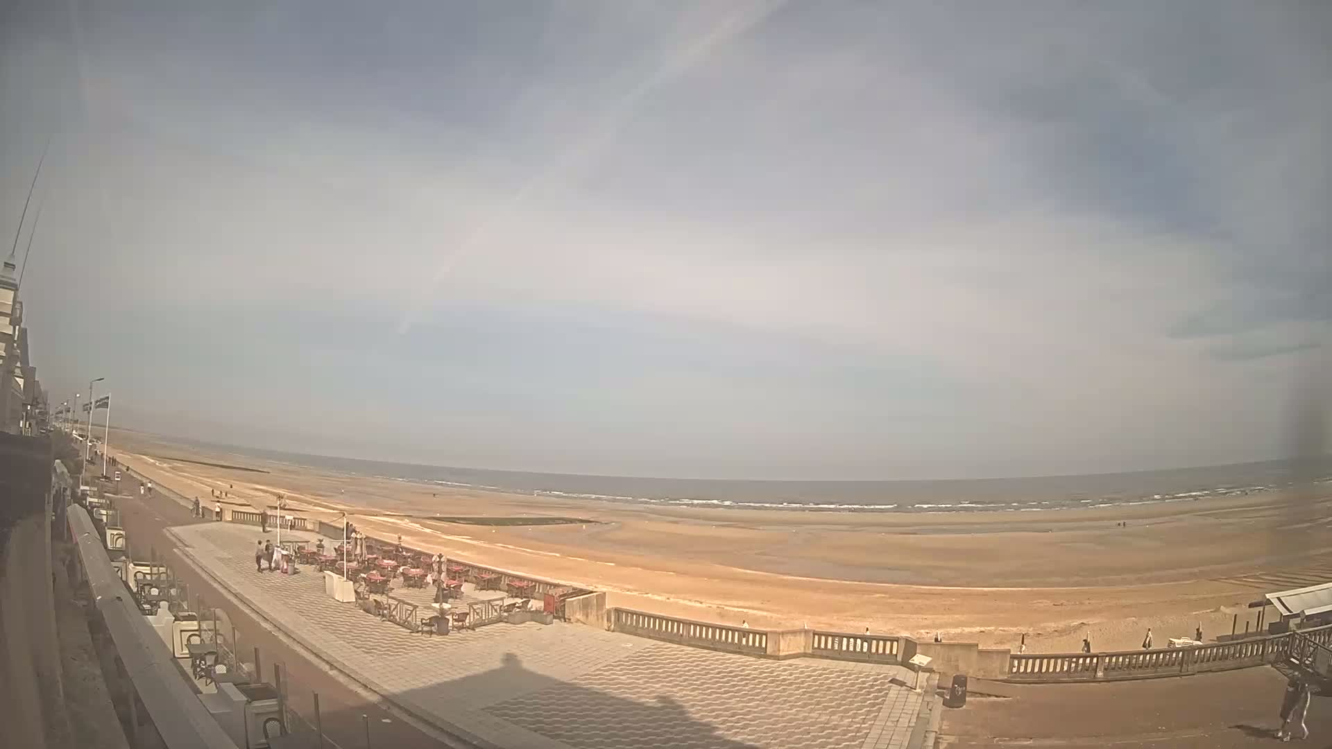 A mostly sunny day reveals a wide sandy beach, a promenade with outdoor seating, and a calm ocean with small waves.