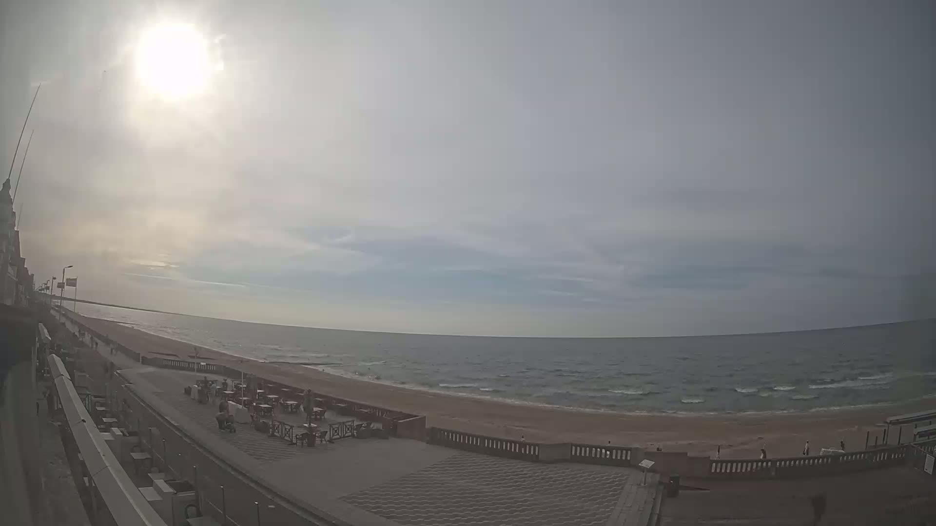 A sunny day at a beach with a partially cloudy sky shows a wide sandy beach with calm waves, an outdoor patio with seating, and a promenade leading to a building.