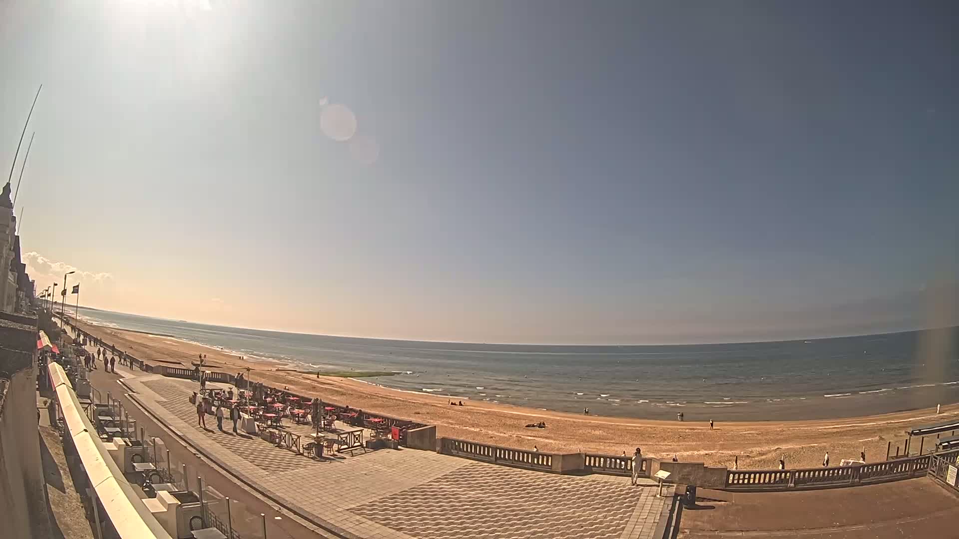 A sunny day at a beachside promenade shows people walking along a paved walkway near an ocean with a sandy beach.
