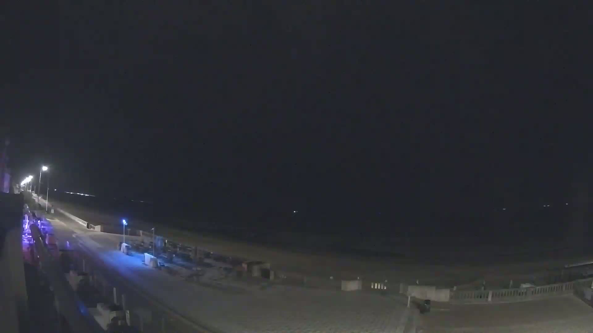 A nighttime view of a mostly empty beach and boardwalk, lit by several streetlights, under a dark sky.
