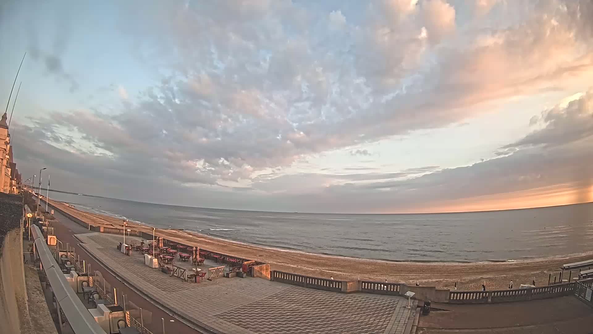 A mostly cloudy sunset illuminates a beach and seaside promenade with outdoor seating areas.