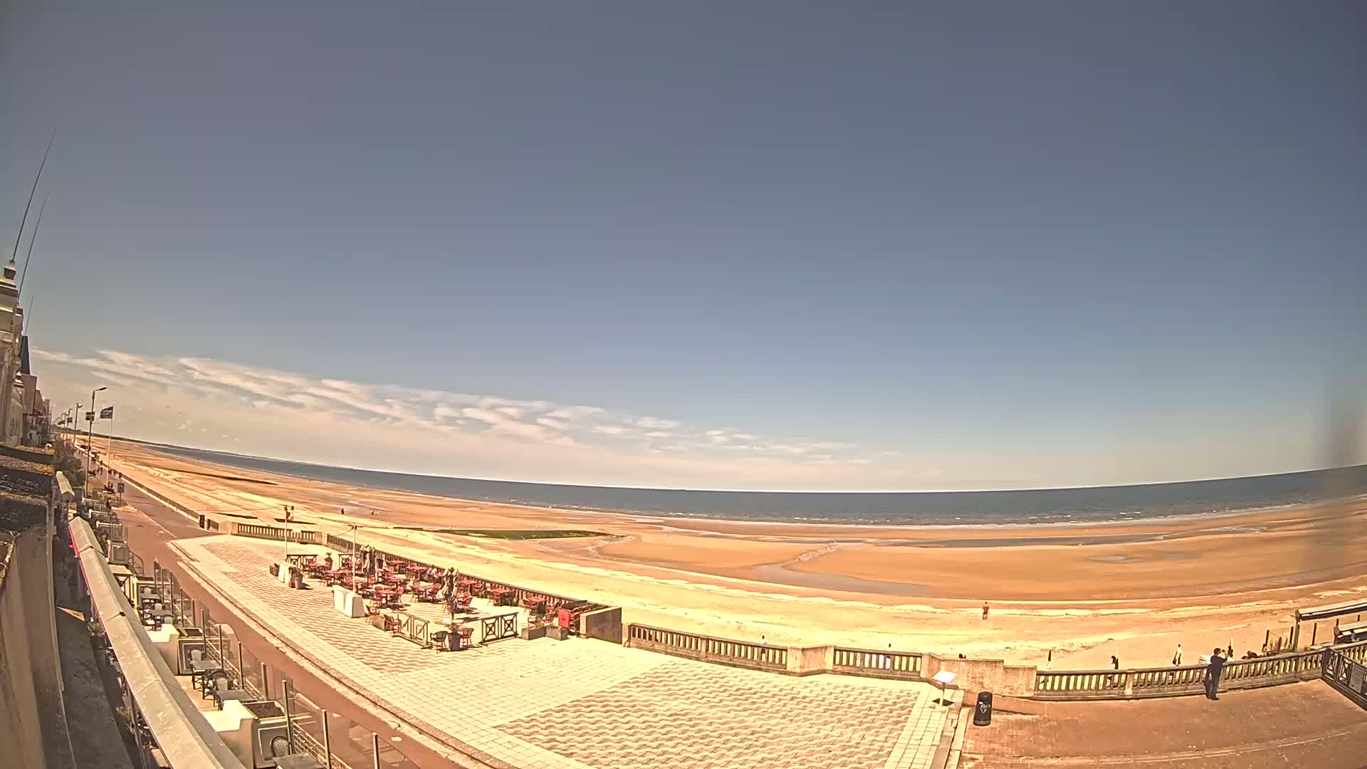 A mostly sunny day reveals a wide sandy beach, a promenade with outdoor seating, and a calm ocean stretching to the horizon.