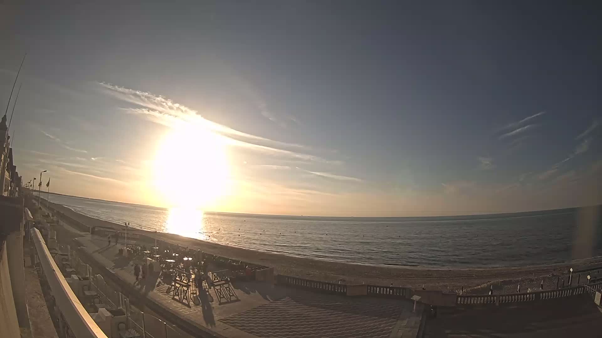 A bright sunset over a calm ocean beach, with people strolling along the promenade near outdoor seating.
