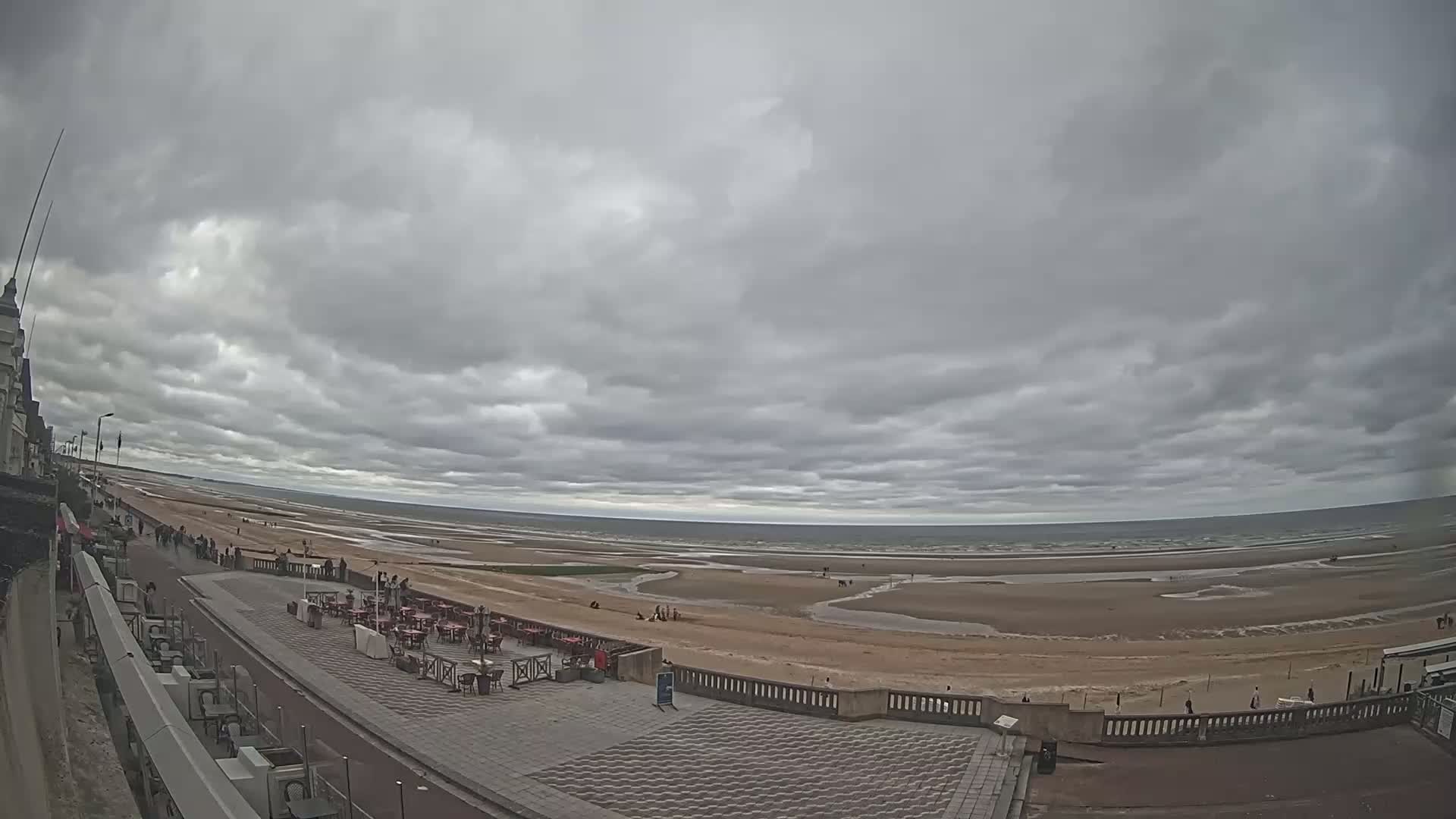 A wide shot shows a mostly overcast sky over a wide, sandy beach with a few people visible, along with an outdoor cafe area and a promenade next to a building.