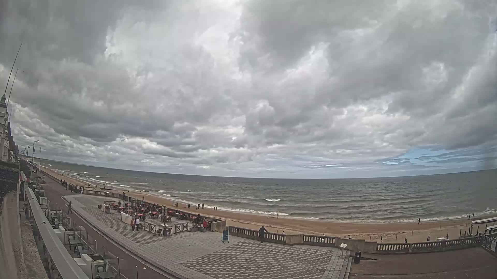 A mostly cloudy day overlooks a beach with a few people walking along the shore and seated at outdoor cafes near the water.