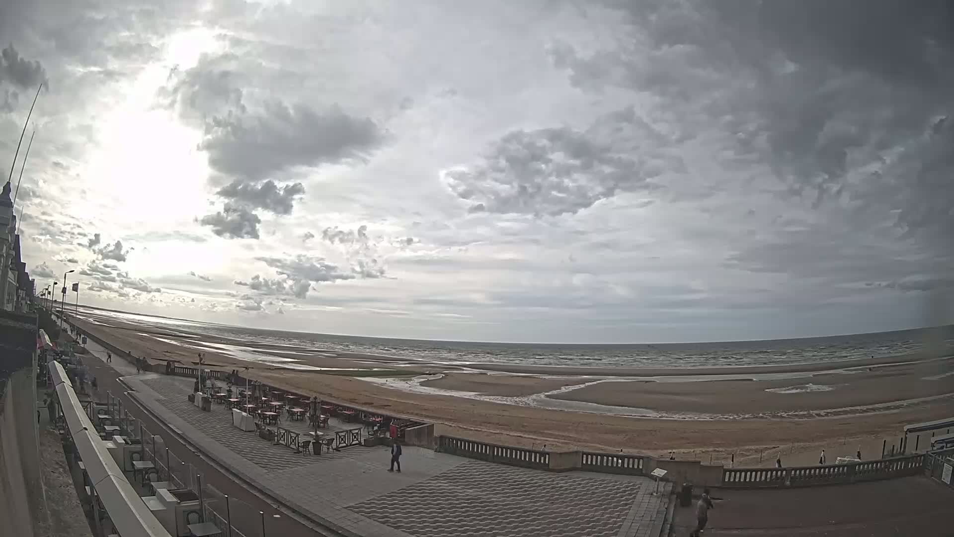A wide shot shows a mostly cloudy sky over a beach with a promenade, outdoor seating areas, and a few people walking.