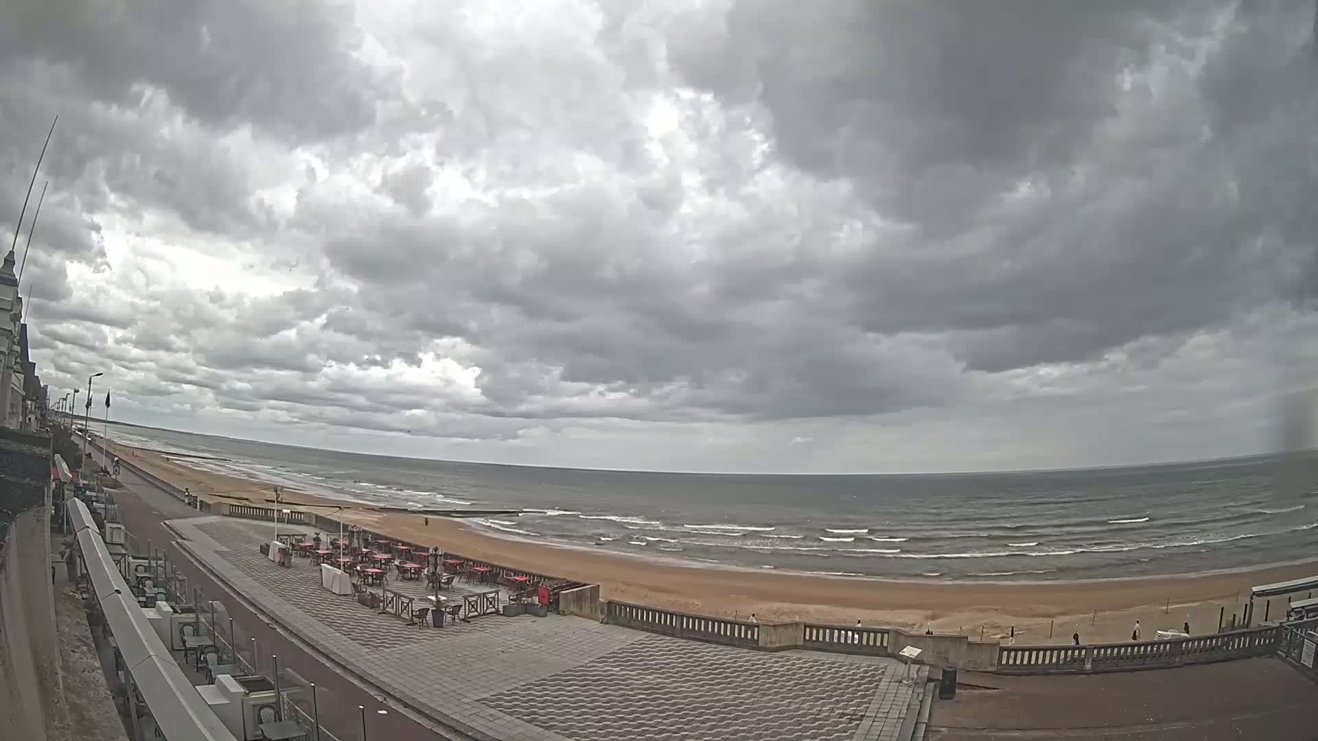 An overcast sky hangs over a beach with moderate waves, a paved walkway, and outdoor seating areas.