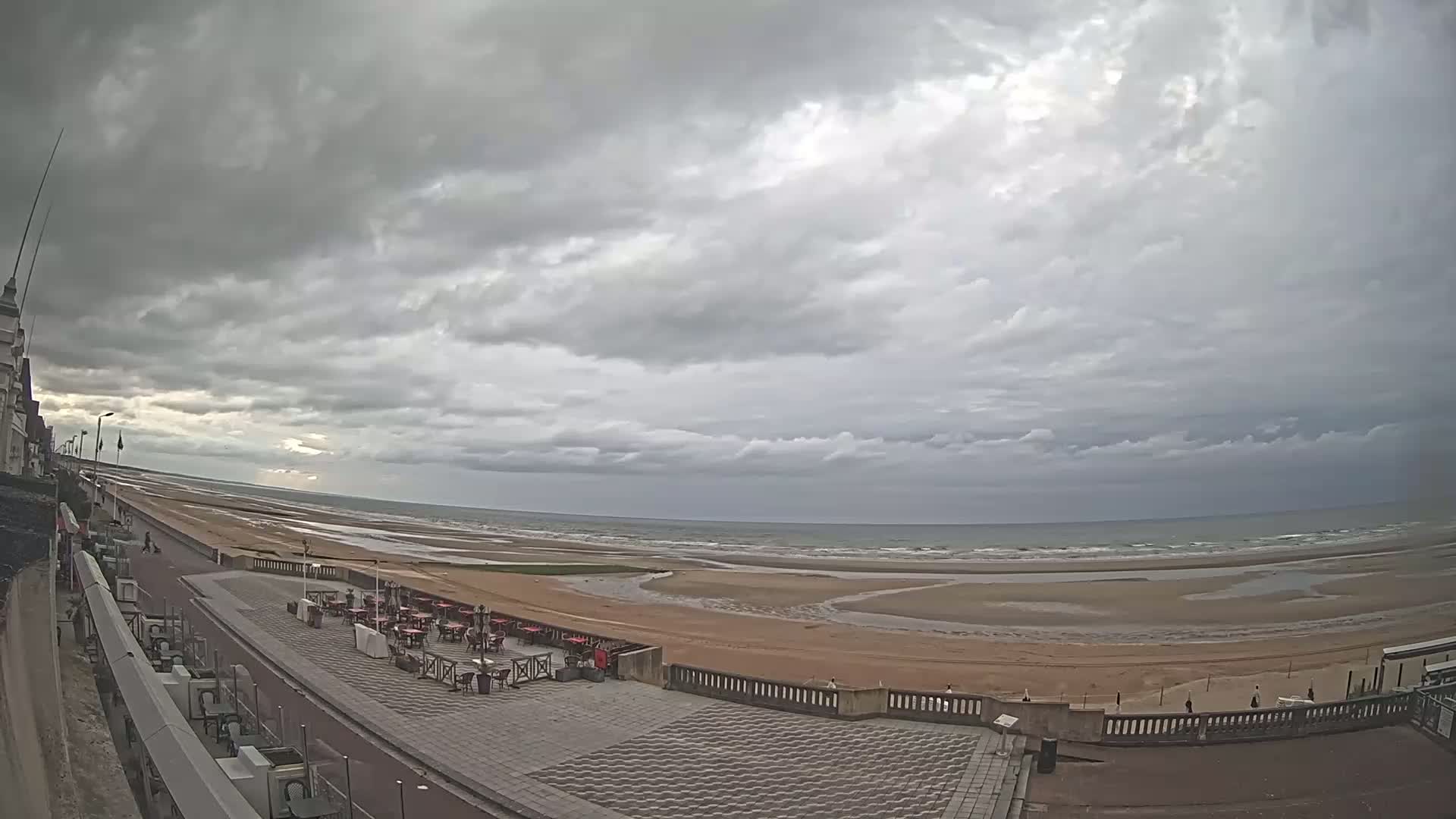 A wide, overcast sky looms over a mostly empty beach with a paved walkway and outdoor seating area next to a building.