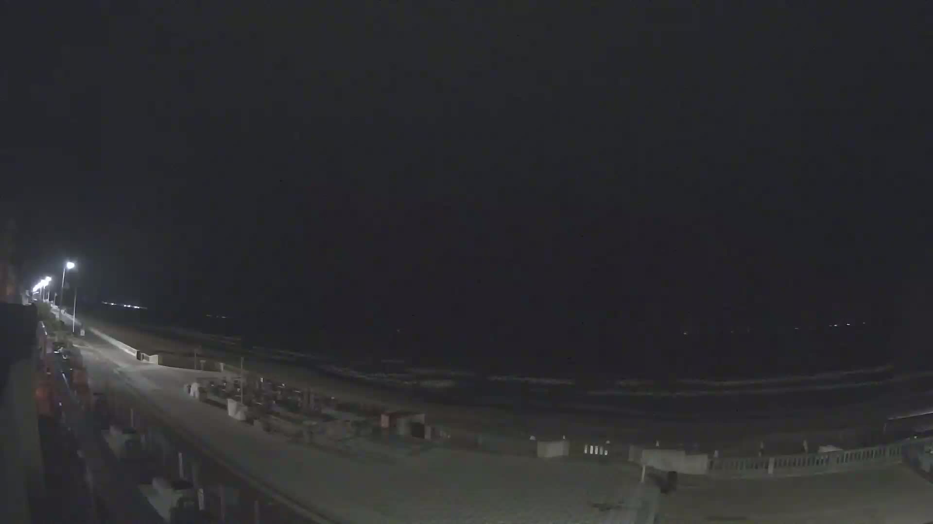 A nighttime view of a beach with a paved walkway lined with lights, showing a dark ocean and a mostly empty beach.