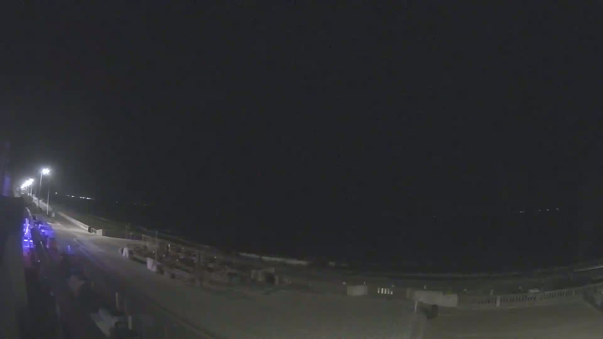 A nighttime view of a mostly empty boardwalk and beach, illuminated by streetlights, under a dark, clear sky.