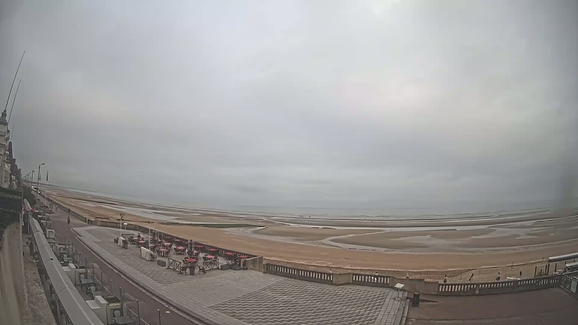A wide, overcast view shows a paved beachfront promenade with an outdoor restaurant area overlooking a wide expanse of sandy beach and calm sea.