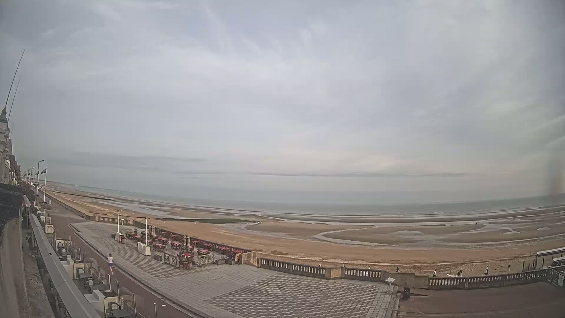 A wide, overcast view shows a paved seaside promenade with outdoor seating areas overlooking a wide expanse of sandy beach and calm ocean.