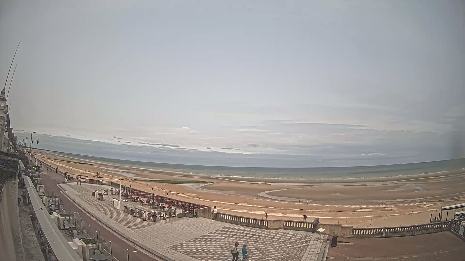 An overcast day reveals a wide expanse of sandy beach and calm ocean, overlooked by a paved promenade with outdoor seating and a few people walking.