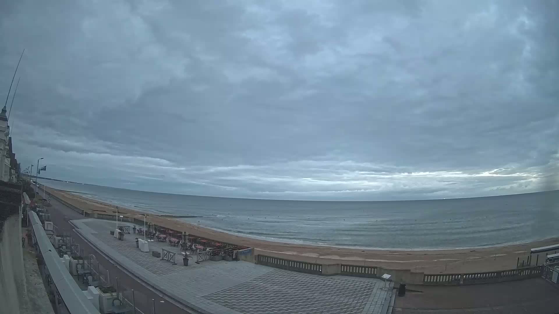 A mostly cloudy sky hangs over a calm ocean beach with outdoor seating areas and a paved walkway.
