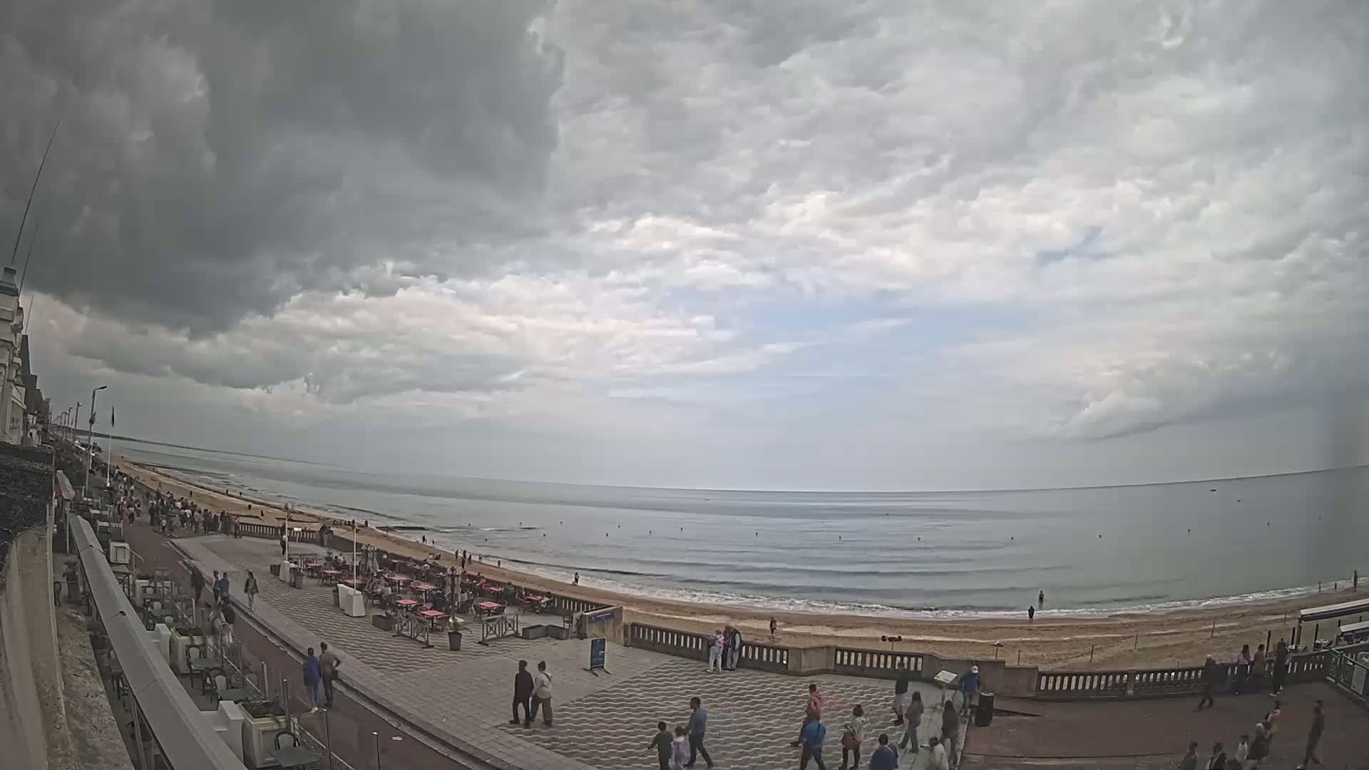 A mostly cloudy day shows an ocean beach with people walking along a paved promenade lined with outdoor seating.