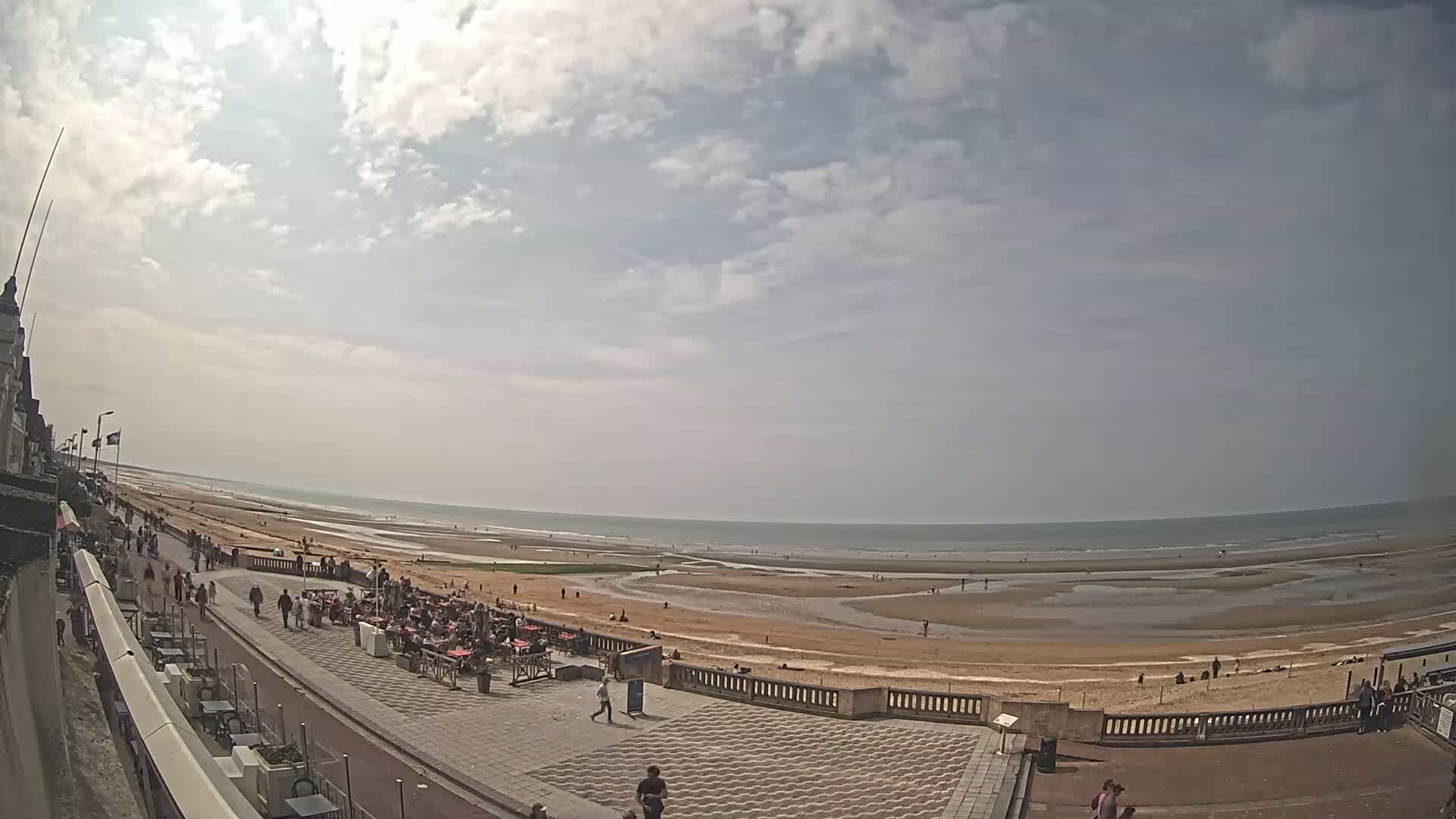 A partly sunny day at a beach shows people strolling along a boardwalk near the ocean, with several outdoor cafes set up on the promenade.
