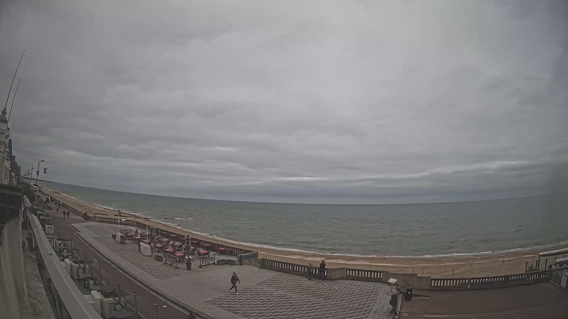 A mostly overcast sky hangs over a calm sea with a sandy beach and paved promenade featuring several outdoor cafe seating areas and a few pedestrians.