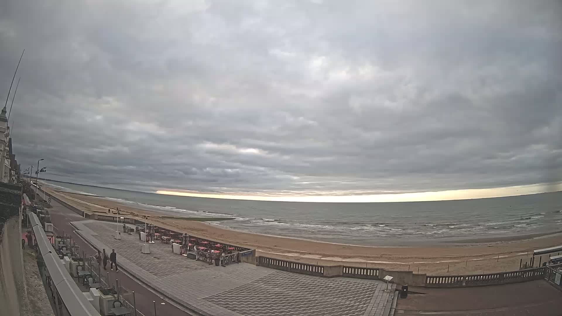A mostly cloudy day overlooks a beach with a promenade, several people walking, and outdoor seating areas along the coast.