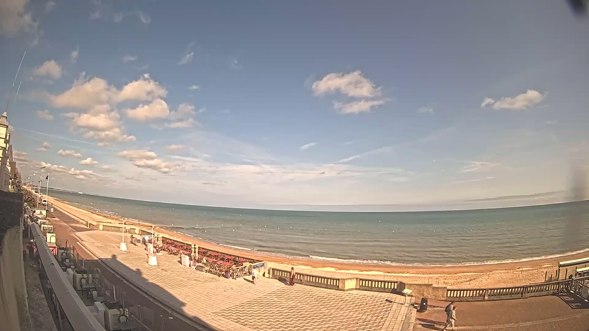 A mostly sunny day shows a wide, sandy beach with calm ocean water, a paved beachfront promenade with outdoor seating areas, and a few people walking along the shoreline.