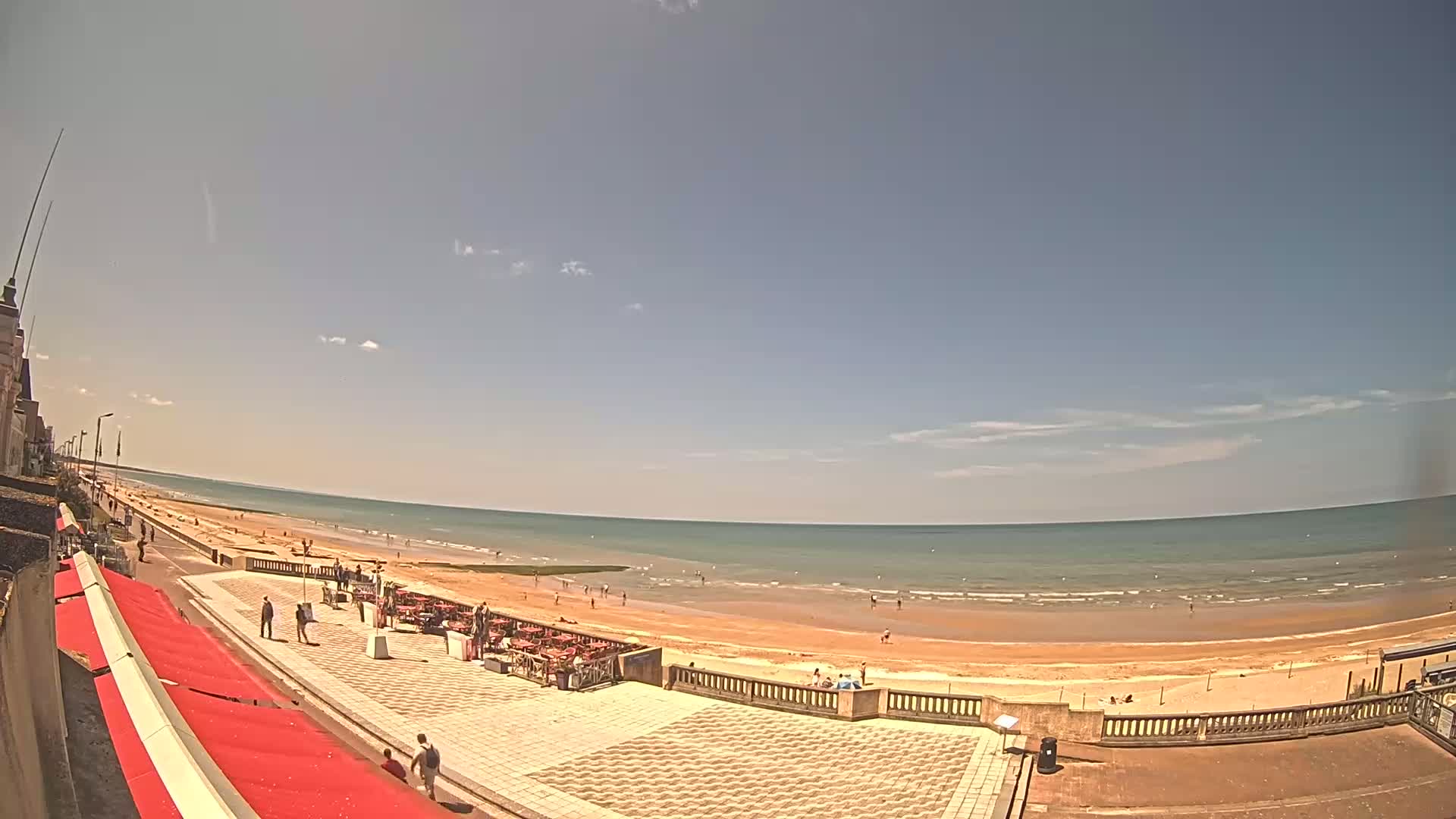 A sunny day at a beach shows a wide sandy shore, calm ocean waves, and a paved promenade with some people and outdoor seating.
