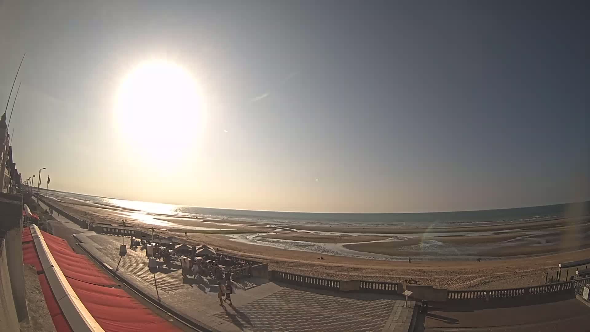 A sunny day at a beach shows a wide sandy shore, calm ocean, and a boardwalk with outdoor seating areas.