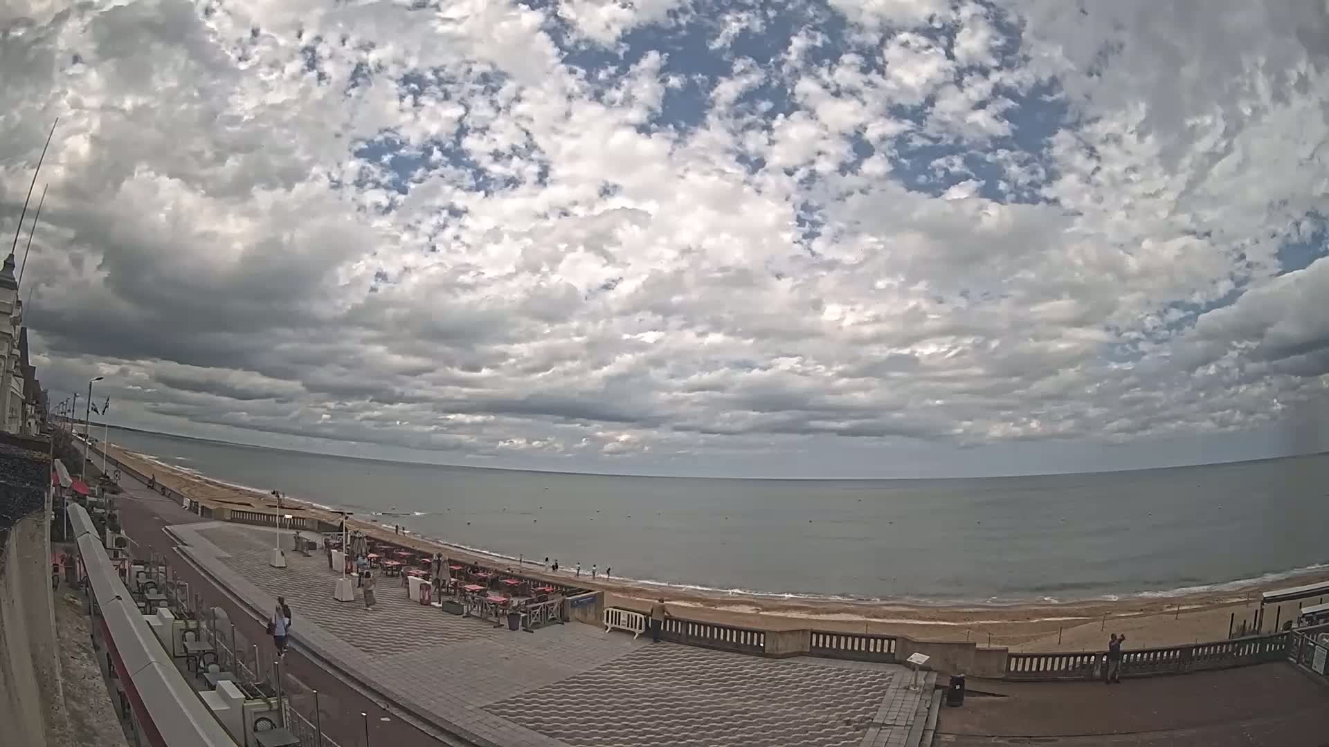 A mostly cloudy sky overlooks a beach with several people and outdoor seating areas along a paved walkway next to a building.
