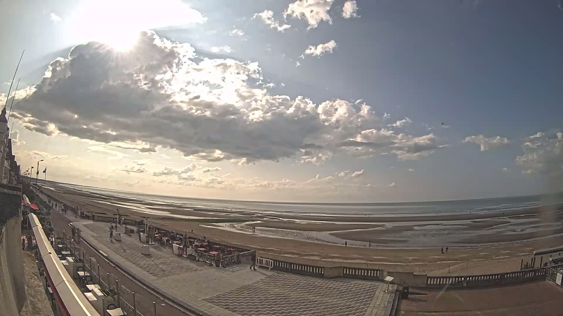 A partly sunny day at a beach with a wide expanse of sand and shallow water at low tide, viewed from an elevated position overlooking a promenade with outdoor seating.