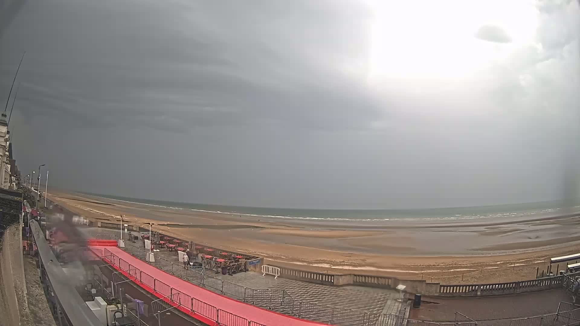 A wide shot shows a mostly overcast sky over a beach with a red carpet leading to some outdoor seating under a stormy sky.