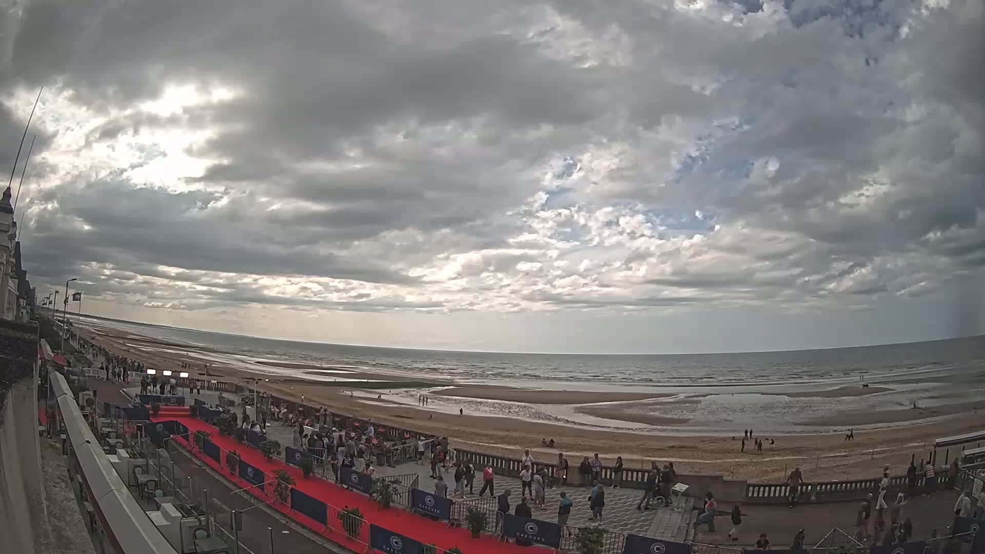 A mostly cloudy sky overlooks a wide sandy beach with a calm ocean and a red carpet leading to a crowd of people near a building.