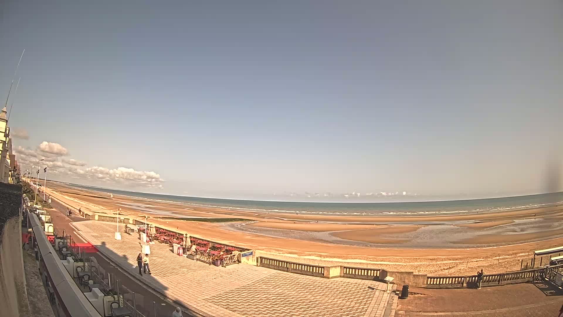 A mostly sunny day reveals a wide sandy beach, a paved promenade with outdoor seating, and a building at the edge of the frame.