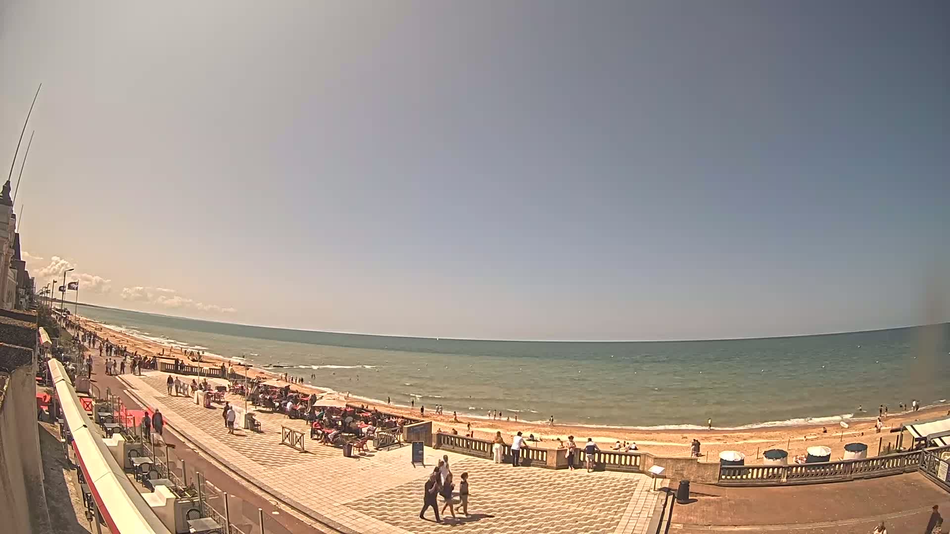 A sunny day at a beachside promenade shows people walking near cafes and restaurants overlooking a calm ocean with a sandy beach.