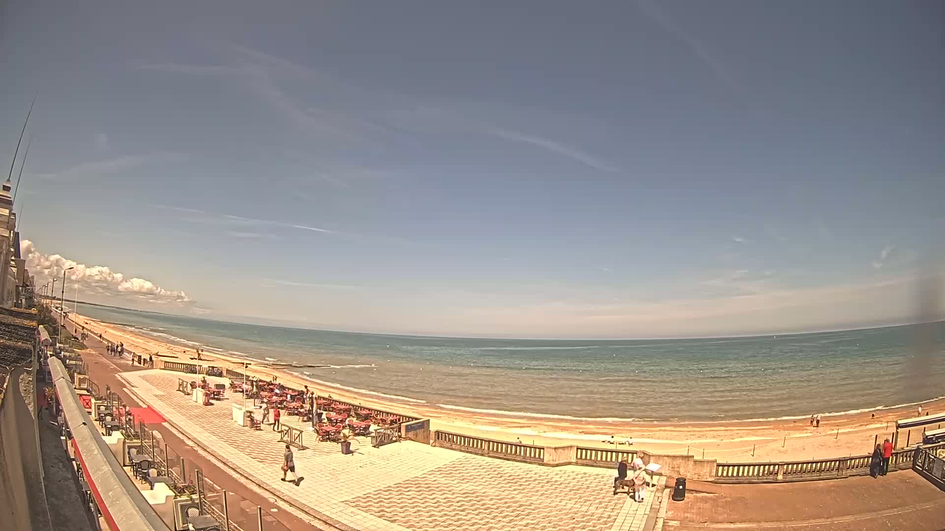 A mostly sunny day at a beachside promenade shows people walking along a sandy beach and sitting at outdoor tables at cafes.