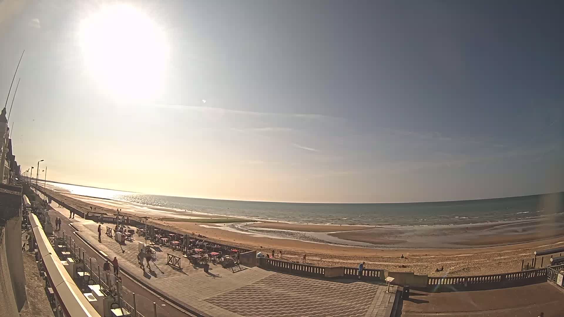 A sunny day at a beachside promenade shows people walking and sitting at outdoor cafes, overlooking a wide sandy beach and calm ocean under a bright sun.