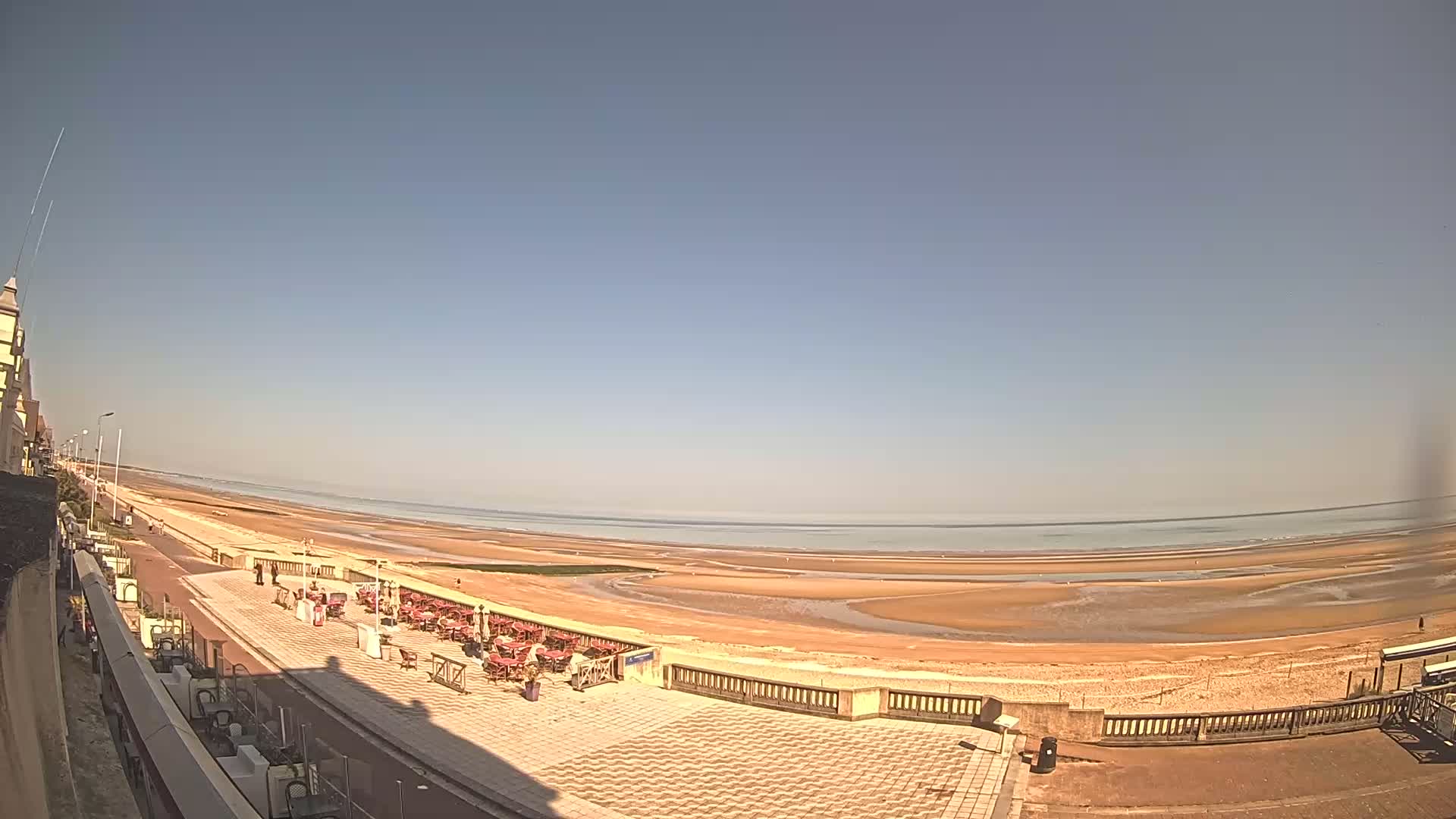 A paved seaside promenade with outdoor seating overlooks a wide, sandy beach and calm ocean under a clear blue sky.