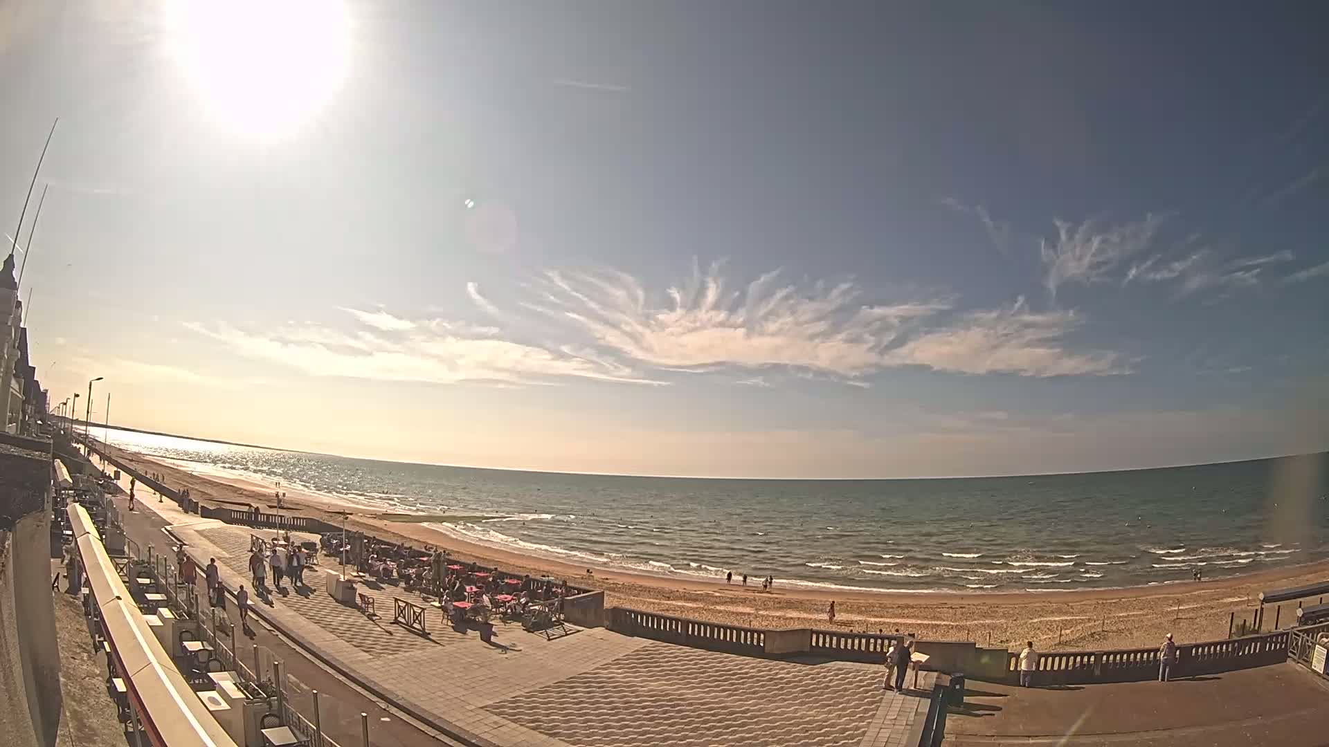 A sunny day at a beach shows a sandy shoreline, calm ocean waves, and a promenade with people walking and sitting at outdoor cafes under a partly cloudy sky with unusual cloud formations.