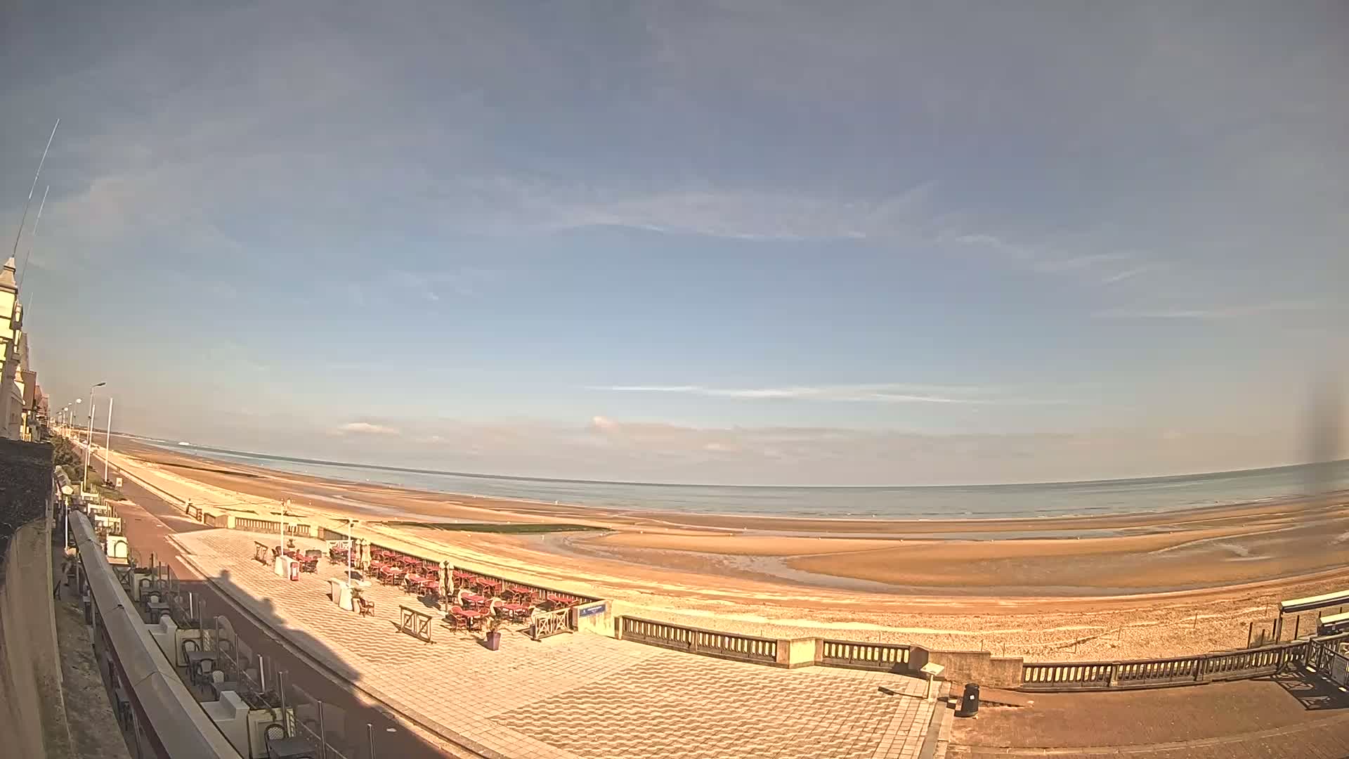 A wide, sandy beach stretches along a calm ocean under a mostly clear, sunny sky, with outdoor seating areas and a paved walkway visible in the foreground.