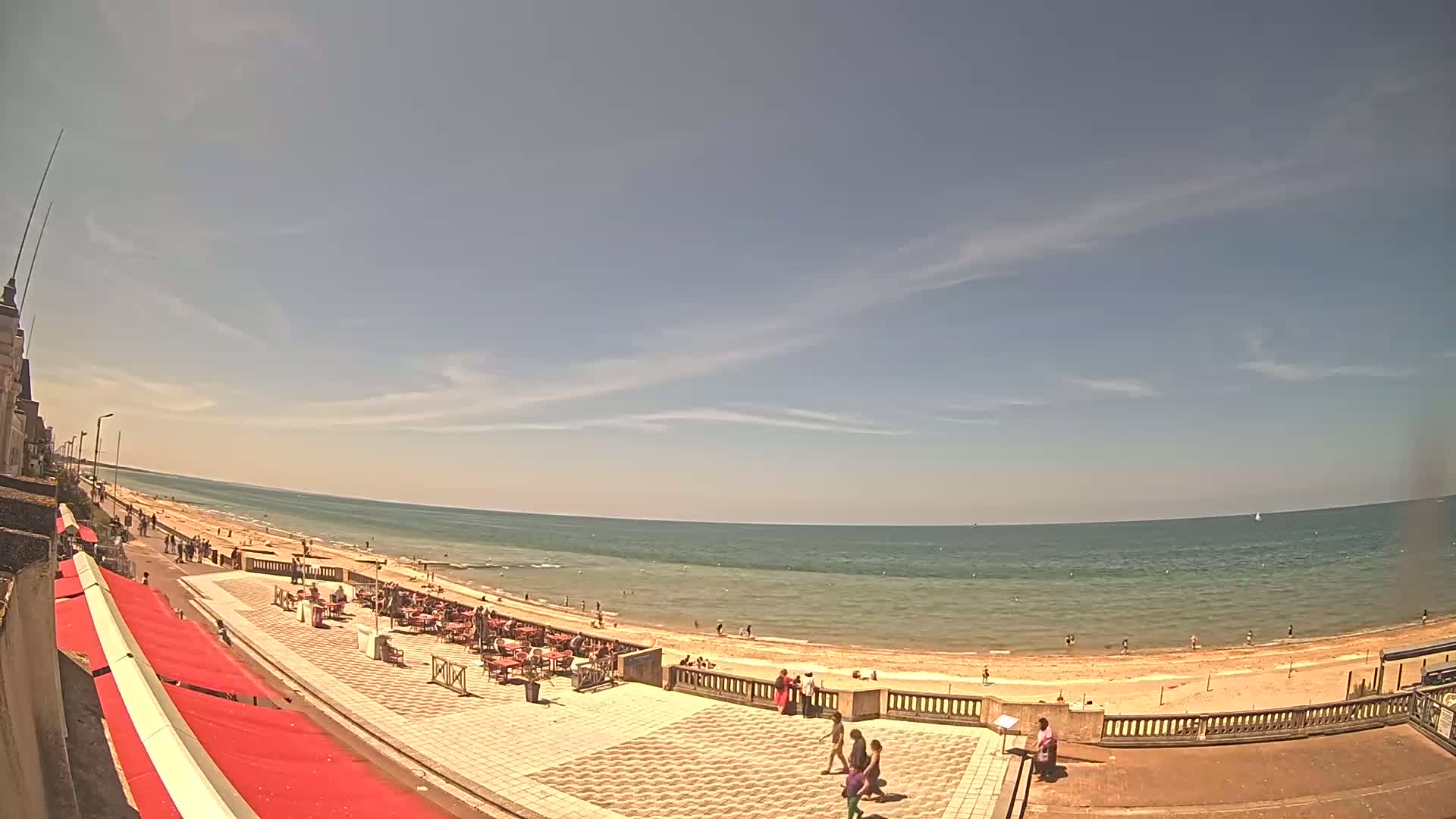 A sandy beach with calm, blue water stretches beneath a mostly clear sky, with a few wispy clouds, and people are enjoying the sunny day on the beach and a paved promenade.