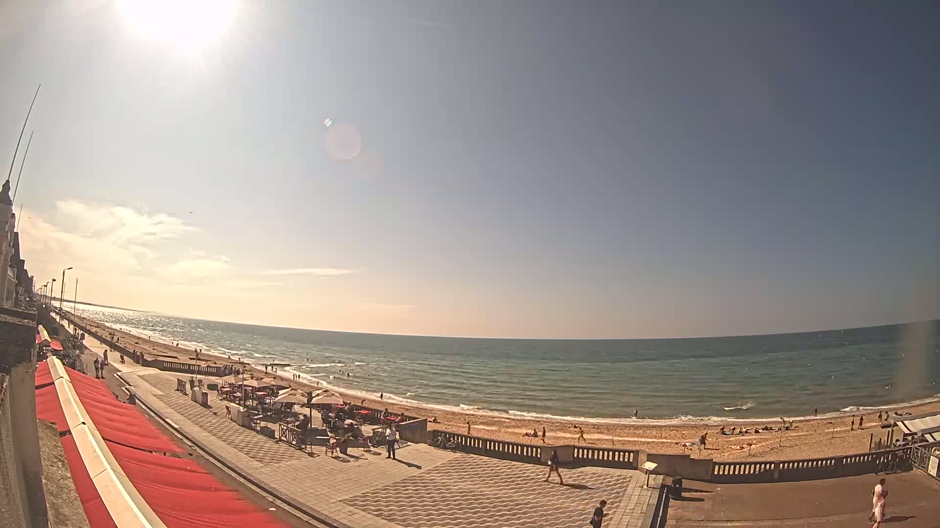 A sunny day at a beach shows a calm ocean, sandy shore with scattered people, and a boardwalk with outdoor seating areas.
