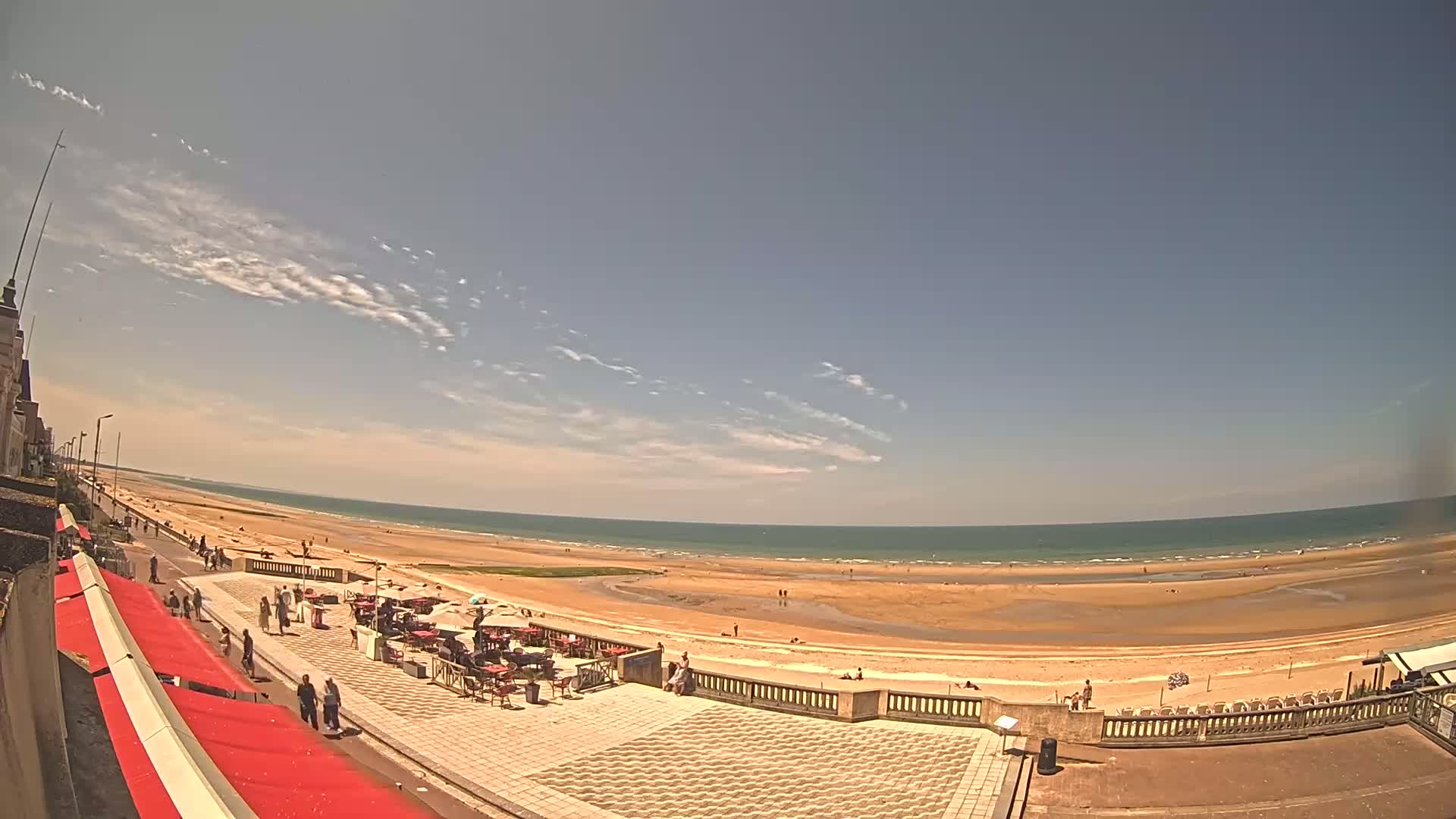 A sandy beach with a calm ocean stretches under a mostly sunny sky with thin, wispy clouds, as people stroll along a paved beachfront promenade lined with outdoor seating.
