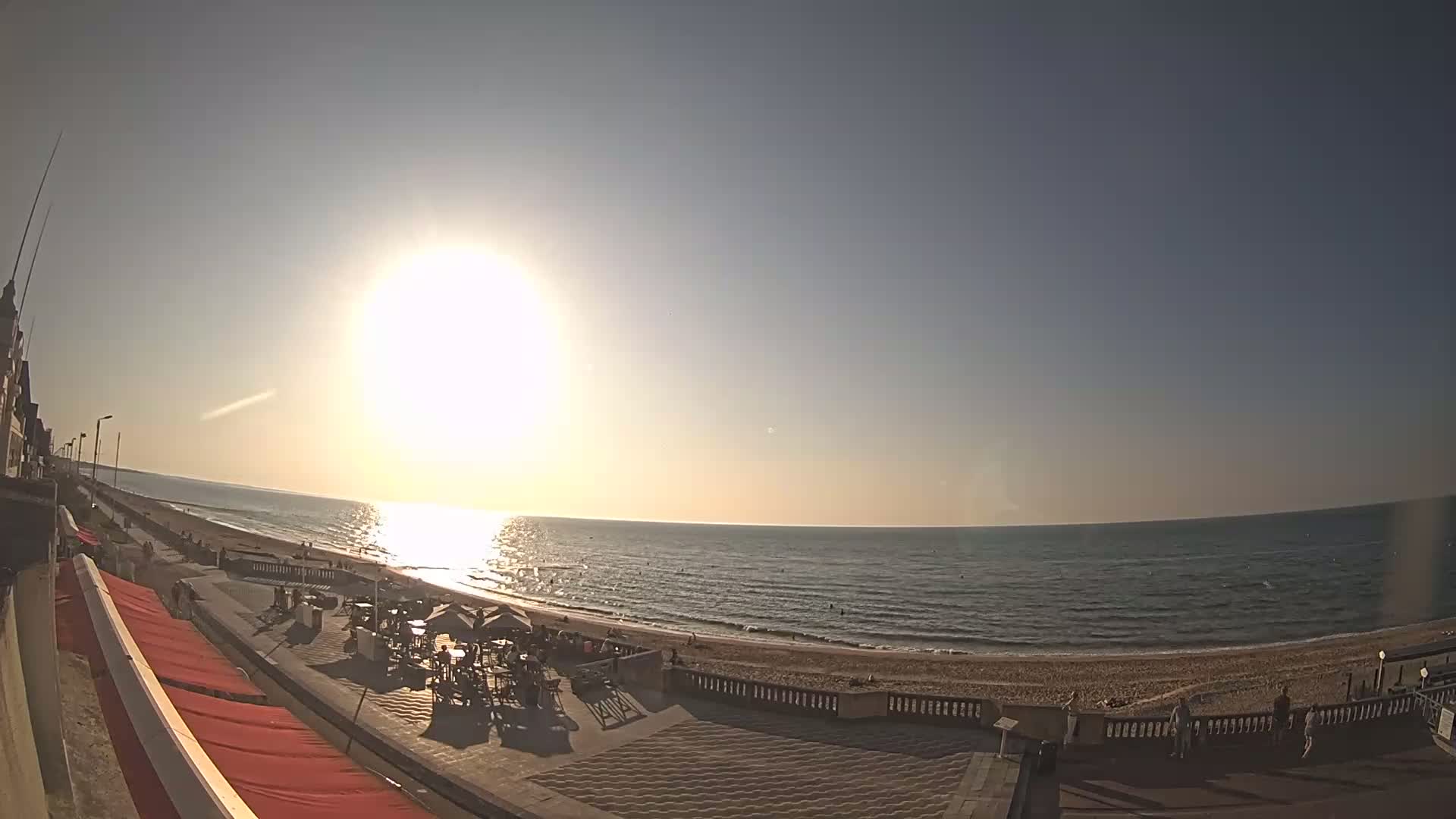A bright sun shines over a calm ocean beach, where people relax at outdoor cafes and stroll along a promenade.