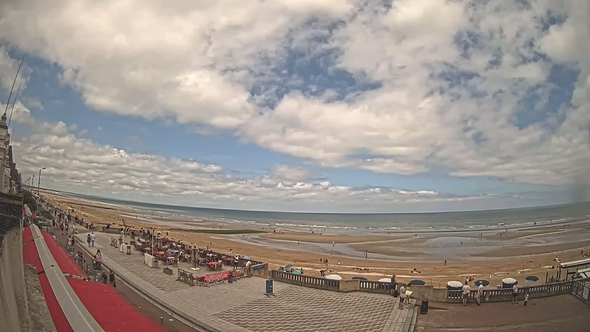 A partly cloudy day at a beach with people sitting at outdoor cafes and walking along a paved walkway overlooking the sand and ocean.