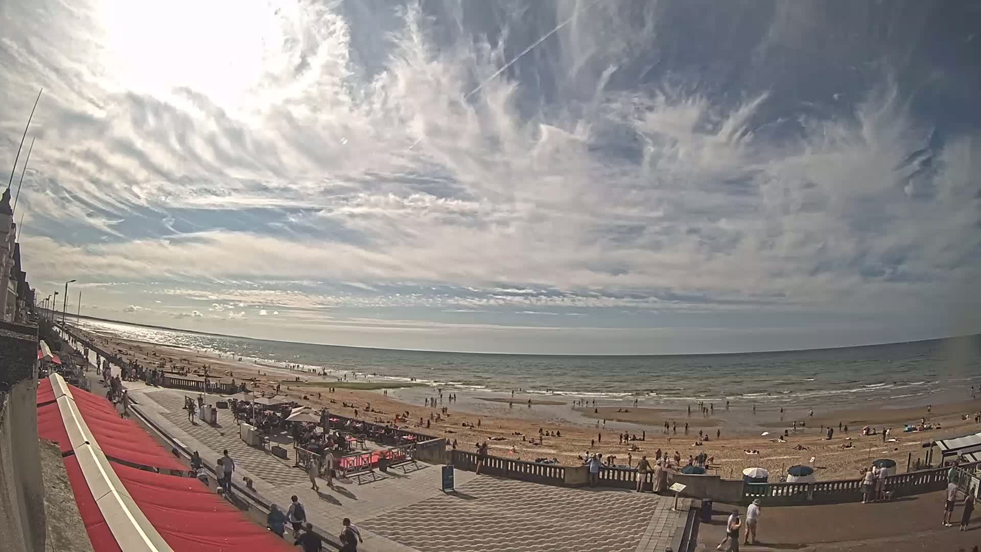 A sunny day at a beach shows a wide sandy shore with many people, a promenade with outdoor seating, and a partly cloudy sky filled with wispy cirrus clouds.