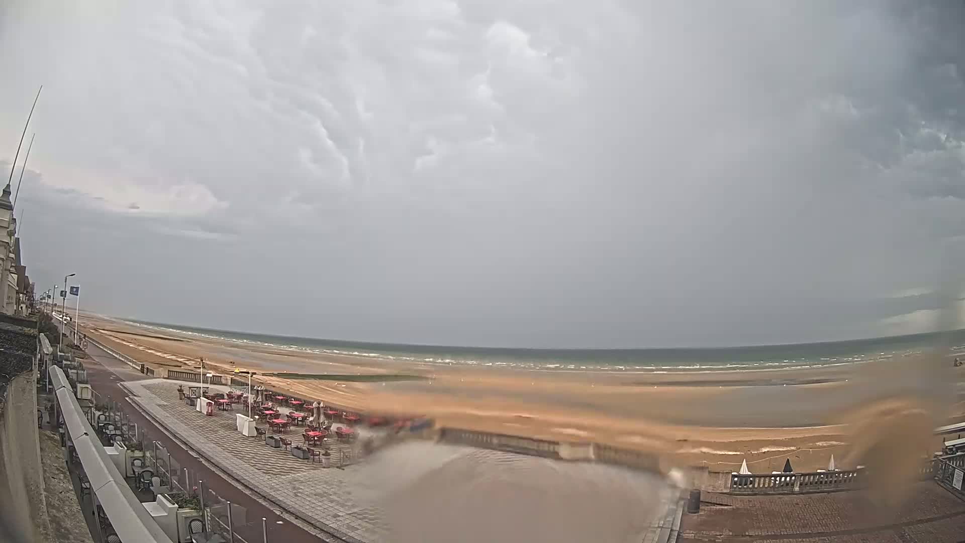 A wide shot of an overcast beach scene shows a sandy beach with a calm sea, an outdoor patio with red tables and chairs, and a paved walkway leading to a building.