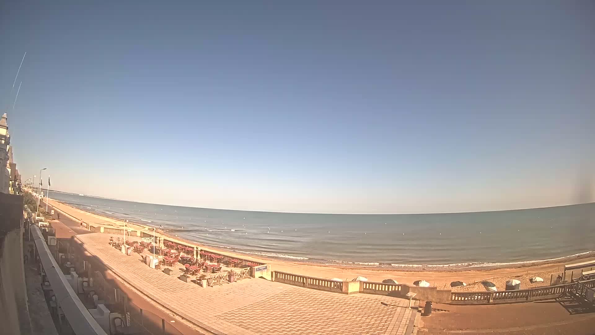 A seaside promenade with an outdoor cafe overlooks a calm, sunny ocean beach.
