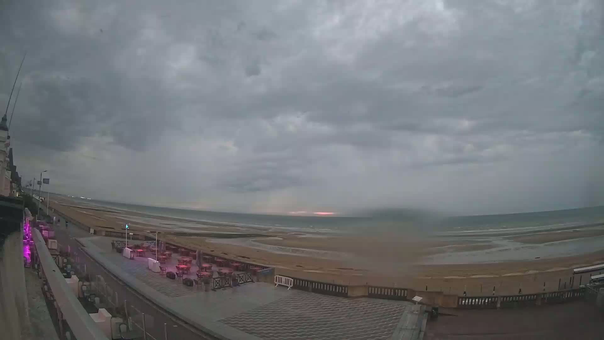 A mostly cloudy, overcast sky hangs over a sandy beach with a calm sea, and outdoor seating at a beachfront restaurant.