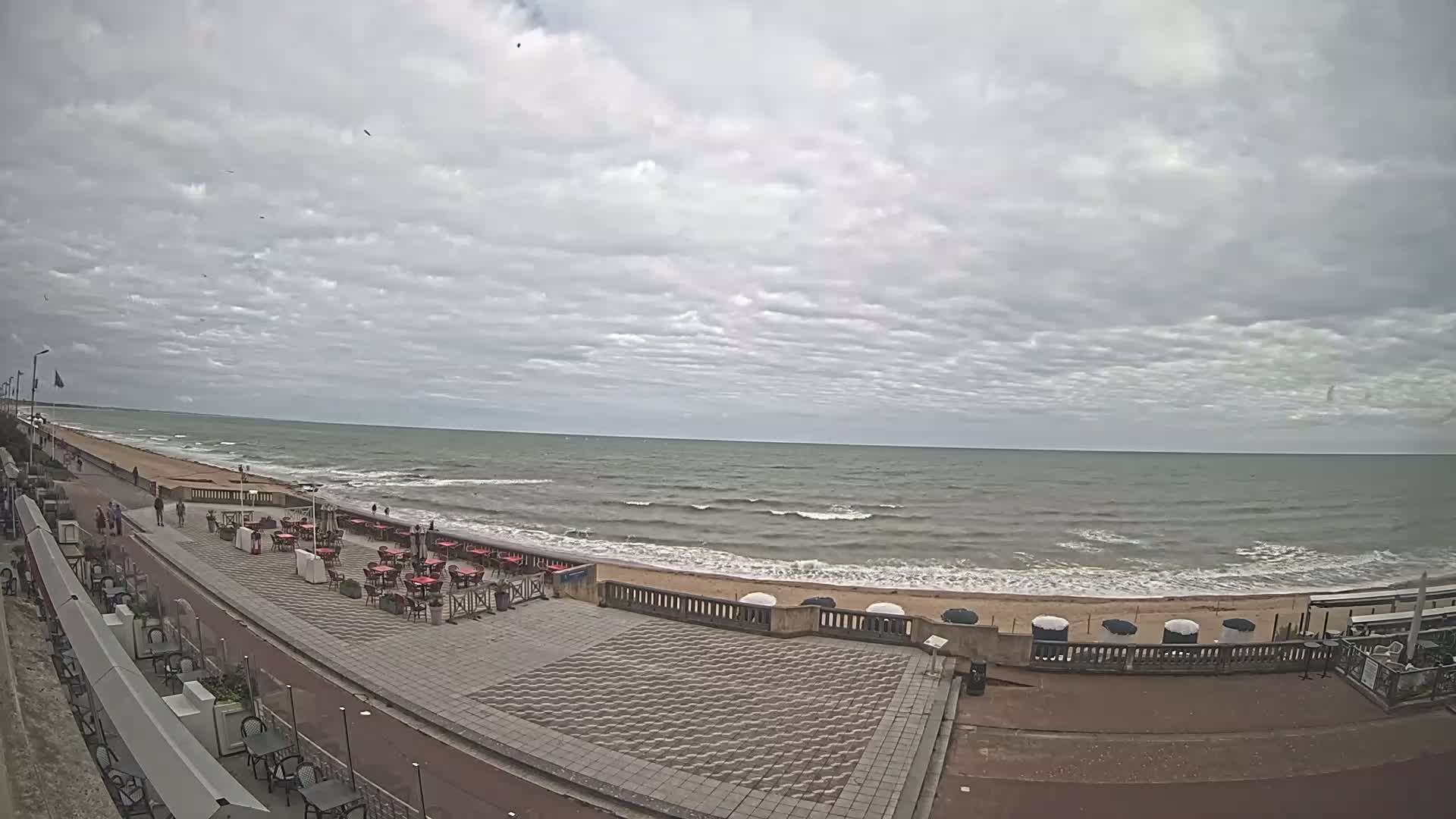 A paved beachfront promenade with outdoor seating overlooks a wavy ocean under a cloudy sky.