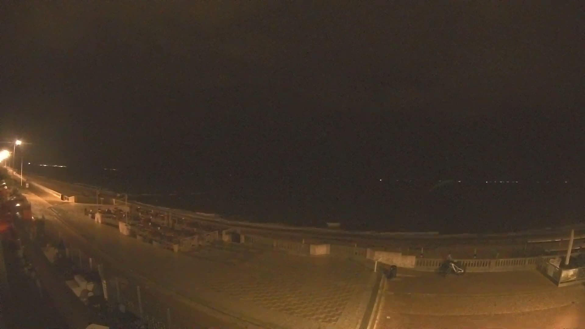 A paved beachfront promenade with outdoor seating overlooks a wavy ocean under a cloudy sky.