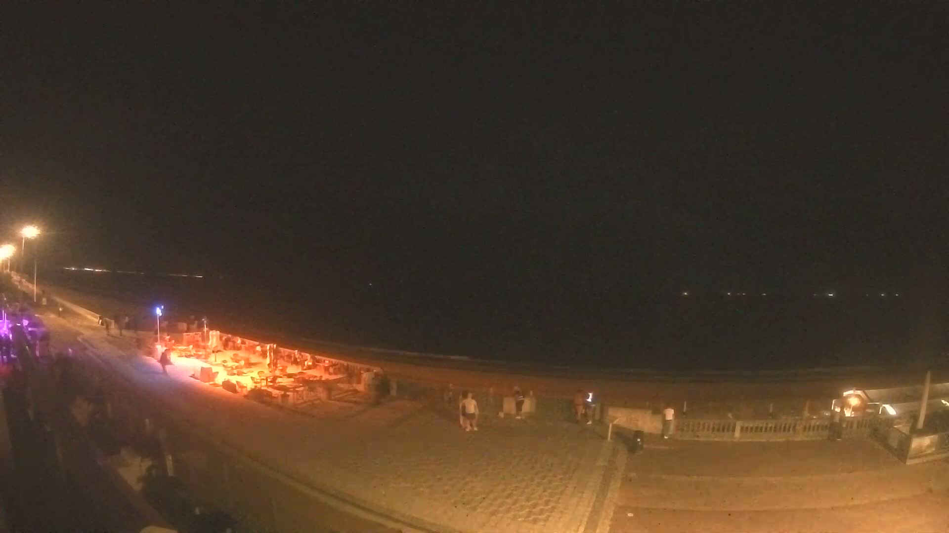 A nighttime beach scene shows people gathered near an illuminated area on a paved walkway next to a dark ocean under a clear sky.
