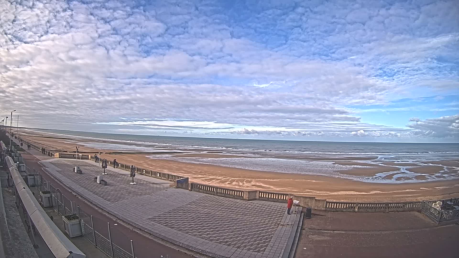 A nighttime beach scene shows people gathered near an illuminated area on a paved walkway next to a dark ocean under a clear sky.
