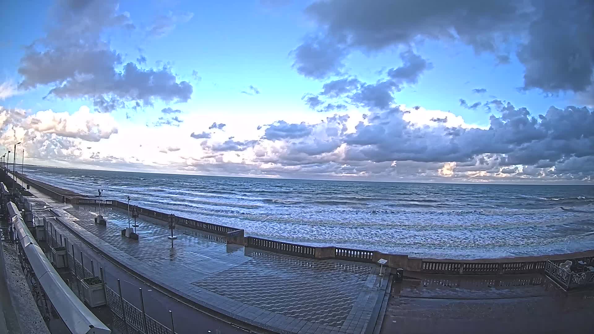 A wide, wet promenade lines a choppy ocean under a dynamic sky with scattered clouds and occasional bright patches of sunlight.